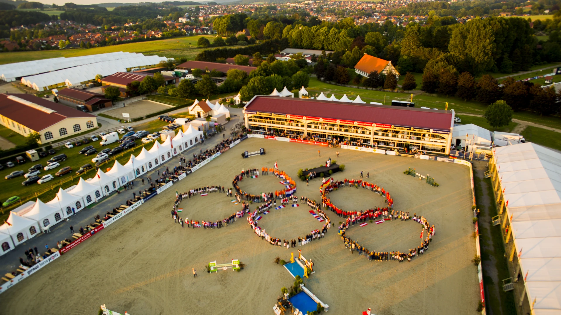 Luftaufnahme eines Pferdeparcours mit Menschen, die olympische Ringe im Sand formen.