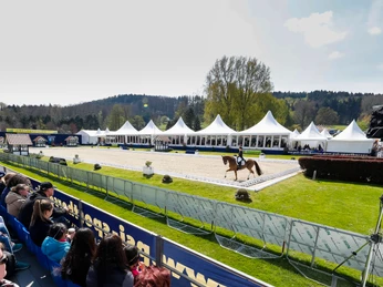 Reitturnier mit Zuschauern, weiße Zelte im Hintergrund, Pferd und Reiter auf Sandplatz.