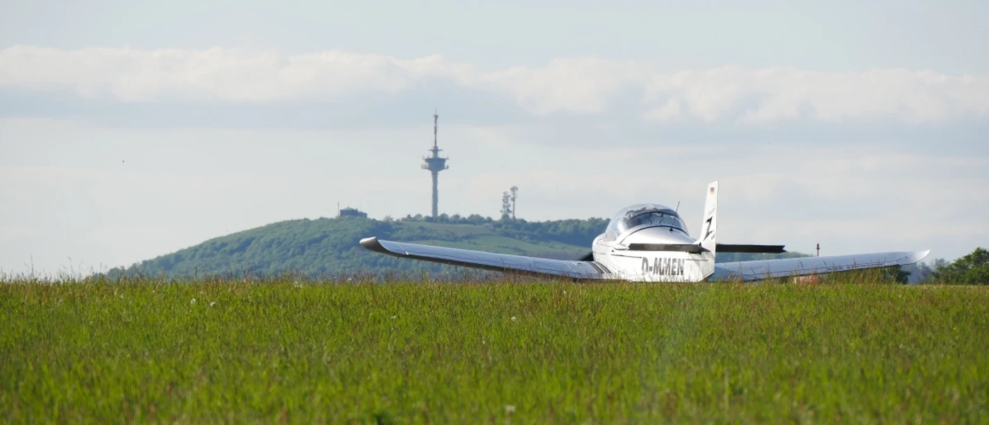 Ein kleines Flugzeug steht auf einer grünen Wiese mit dem bewaldeten Köterberg und einem Funkturm im Hintergrund.