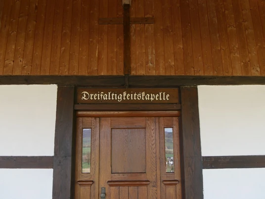 Eingang der Dreifaltigkeitskapelle mit einem Holzkreuz darüber und traditionellem Schriftzug.