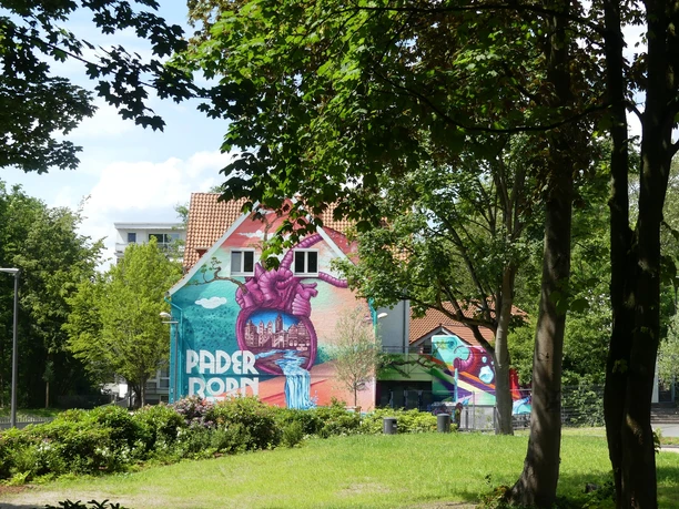 Paderborner Herzgraffiti im Mittleren Paderquellgebiet Graffitihäuserwand in Paderborn: Herzmural mit Landschaftsdetails zwischen Laubbäumen und Wiese.