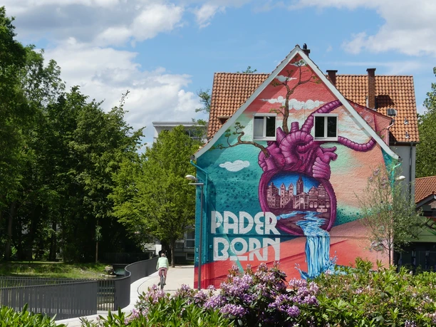 Graffiti-Kunst mit Herz-Motiv und Paderborn-Schriftzug auf einer Hausfassade in grüner Umgebung.
