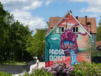 Paderborner Herzgraffiti im Mittleren Paderquellgebiet Graffiti-Kunst mit Herz-Motiv und Paderborn-Schriftzug auf einer Hausfassade in grüner Umgebung.