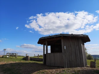 Kleine Holzschutzhütte auf einem Hügel mit weitem Blick über eine Wiesenlandschaft an einem klaren Tag.