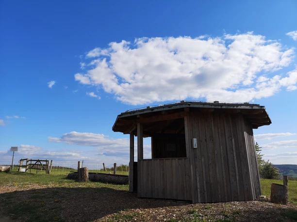Hude-Hütte am Räuschenberg Kleine Holzschutzhütte auf einem Hügel mit weitem Blick über eine Wiesenlandschaft an einem klaren Tag.