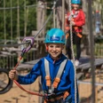 Kind in blauer Kletterausrüstung mit Helm und Sicherheitsseil in einem Abenteuerpark.