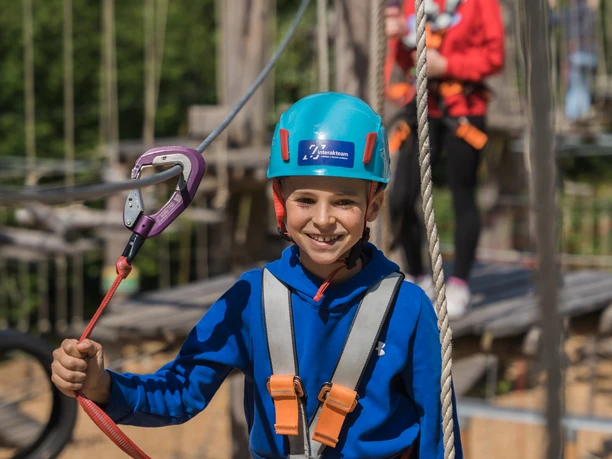 Kind in blauer Kletterausrüstung mit Helm und Sicherheitsseil in einem Abenteuerpark.