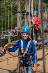 Kletterpark Bielefeld2.jpg Kind in blauer Kletterausrüstung mit Helm und Sicherheitsseil in einem Abenteuerpark.