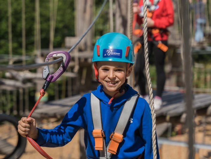 Kletterpark Bielefeld2.jpg Kind in blauer Kletterausrüstung mit Helm und Sicherheitsseil in einem Abenteuerpark.