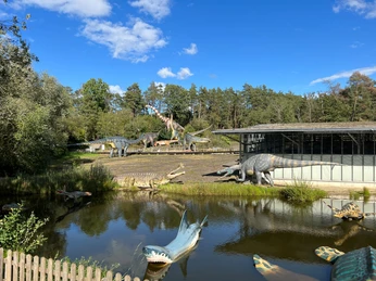 Dinosauriermodelle im Freien, umgeben von Bäumen und Wasserflächen bei blauem Himmel und Sonnenschein.Dinosaur models outdoors, surrounded by trees and bodies of water under a blue sky and sunshine.Dinosaurmodeller i det fri, omgivet af træer og vandområder under en blå himmel og solskin.Dinosaurusmodellen in de open lucht, omringd door bomen en watermassa's onder een blauwe hemel en zonneschijn.