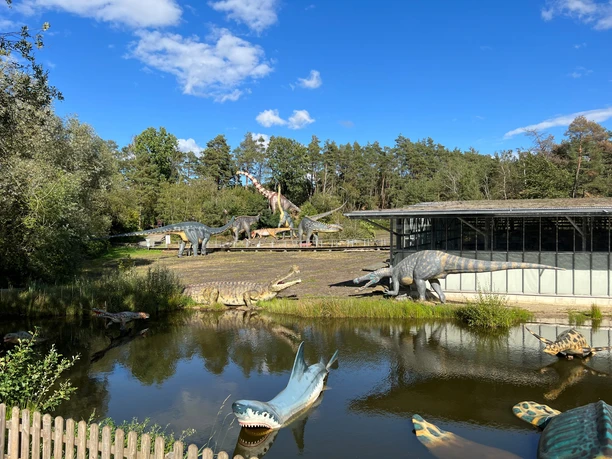 Dinosaur models outdoors, surrounded by trees and bodies of water under a blue sky and sunshine.