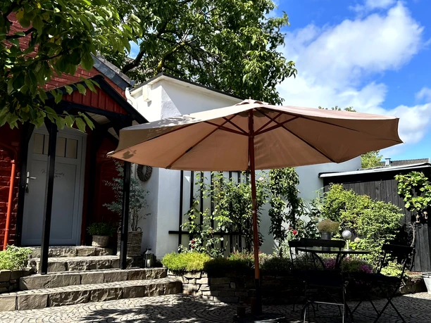 Ferienwohnung "Das rote Ferienhäuschen" Ein sonniger Innenhof mit Terrassenmöbeln und Sonnenschirm, umgeben von grünen Bäumen und blauen Himmel.
