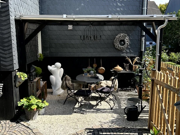 Ferienwohnung "Das rote Ferienhäuschen" Ein gemütlicher, halboffener Patio mit einem runden Tisch, umgeben von Pflanzen und einer Skulptur.