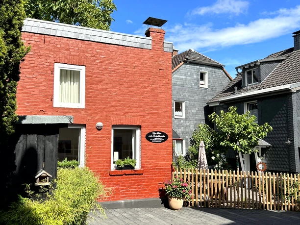 Ferienwohnung "Das rote Ferienhäuschen" Rotes Backsteinhaus mit Holzfensterläden, umgeben von Kopfsteinpflaster, Bäumen und strahlend blauem Himmel.