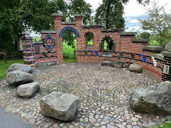 Altenbücken Backsteinskulptur mit bunten Fliesen in runder Anordnung, umgeben von Bäumen und großen Steinen.