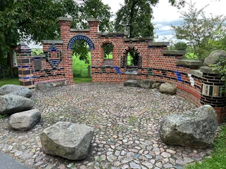 Kreative Backsteinarchitektur mit bunten Fliesen und Natursteinen auf einer runden Pflasterfläche im Grünen.