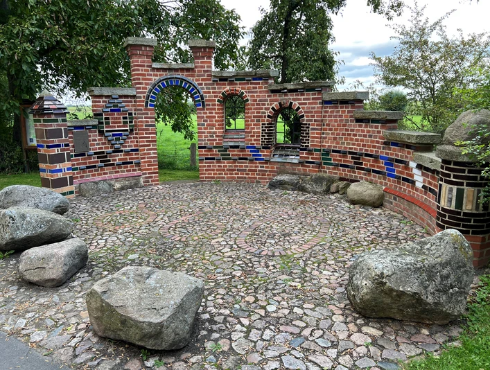 Kreative Backsteinarchitektur mit bunten Fliesen und Natursteinen auf einer runden Pflasterfläche im Grünen.