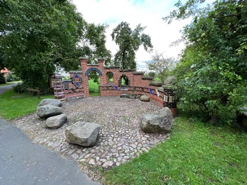 Roter Backsteinbogen mit blauen Details und hölzernen Bänken in einer grünen Parkumgebung.