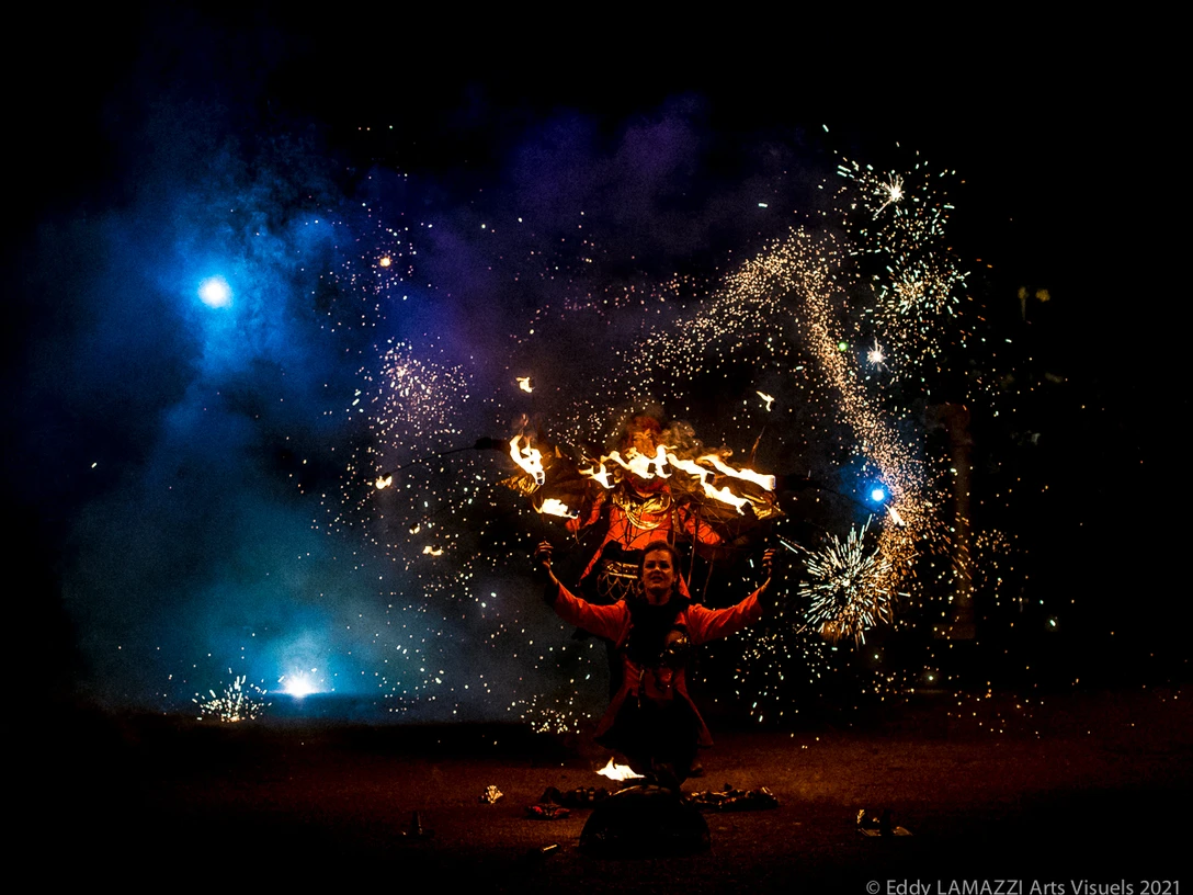 189-Hanabi-circus-©Eddy-LAMAZZI-Arts-Visuels-2021-5962.jpg Feuerkünstler in roter Kleidung mit Funkenregen und blauem Licht hinter ihm, umgeben von Dunkelheit.