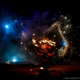189-Hanabi-circus-©Eddy-LAMAZZI-Arts-Visuels-2021-5962.jpg Feuerkünstler in roter Kleidung mit Funkenregen und blauem Licht hinter ihm, umgeben von Dunkelheit.