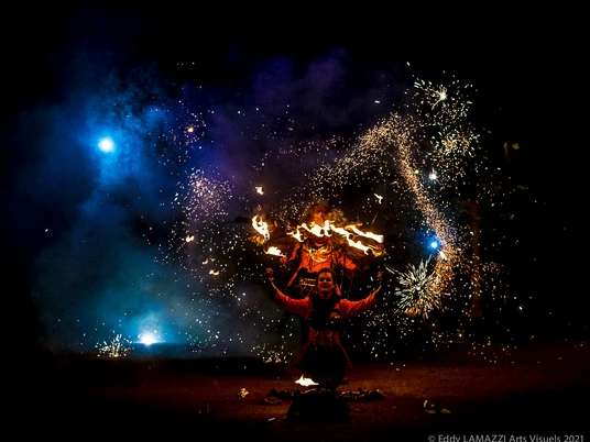 189-Hanabi-circus-©Eddy-LAMAZZI-Arts-Visuels-2021-5962.jpg Feuerkünstler in roter Kleidung mit Funkenregen und blauem Licht hinter ihm, umgeben von Dunkelheit.