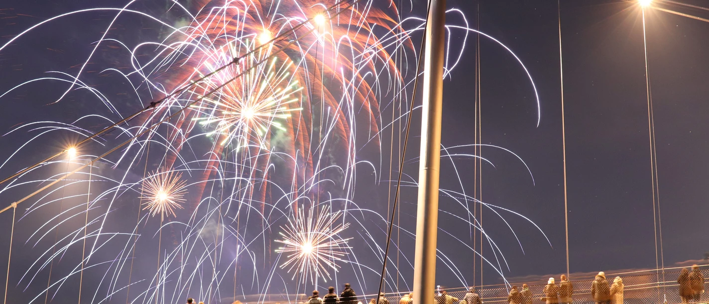 messe_2022_3.JPG Menschenmenge steht auf einer beleuchteten Fußgängerbrücke und beobachtet farbenfrohes Feuerwerk am Nachthimmel.