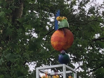 Spatz am Lieblingsort Schutzhütte Apermarsch Eine bunte, fantasievolle Skulptur von Vögeln im Grünen, gekrönt von einem majestätischen Spatz.