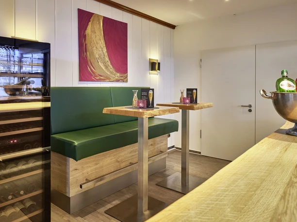 Gemütliche Hotelbar mit grüner Sitzbank, hohen Holztischen und moderner Kunst an der Wand.