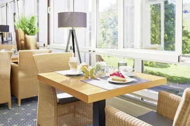 22697_Waldhotel Baerenstein_2023_Kaffee_A.jpg Heller Frühstücksraum mit geflochtenen Stühlen, Tisch mit Kaffee, Croissant und Marmelade, sonniger Ausblick.