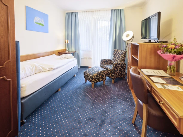 22697_Waldhotel Baerenstein_2023_Zimmer9_01.jpg Geräumiges Hotelzimmer mit Einzelbett, Sessel, Schreibtisch und Flachbildfernseher, freundlich beleuchtet.