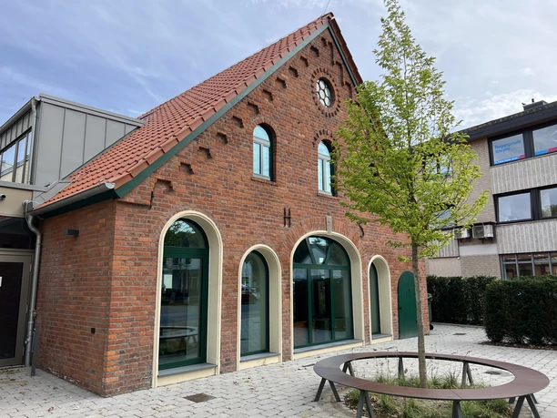 The Leeste Cultural Barn, with its red-tiled roof, round arches, and clock tower, set against a small tree.