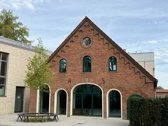 Backsteingebäude mit Giebel, Sprossenfenstern und Rundbögen, angelehnt an einen modernen Anbau.