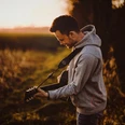 Kevin Guennoc Ein Mann spielt Gitarre auf einem Feldweg bei Sonnenuntergang, umgeben von herbstlicher Natur.