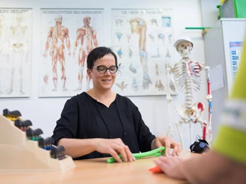 221021-FotoChristianSchwier-0304.jpg Eine Frau trainiert mit einem elastischen Übungsband in einer Physiotherapiepraxis, umgeben von anatomischen Postern und einem Skelettmodell.