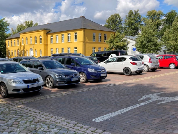 Parkplatz_Rathaus1_22082023_PetraRoemer.jpg Parkplatz mit Autos. Im Hintergrund steht das gelbe Rathaus