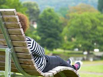 Park Klinik Bank_©Sascha Reichert-2928.jpg Person sitzt entspannt auf einer geschwungenen Holzbank in einem grünen Park mit Bäumen im Hintergrund.