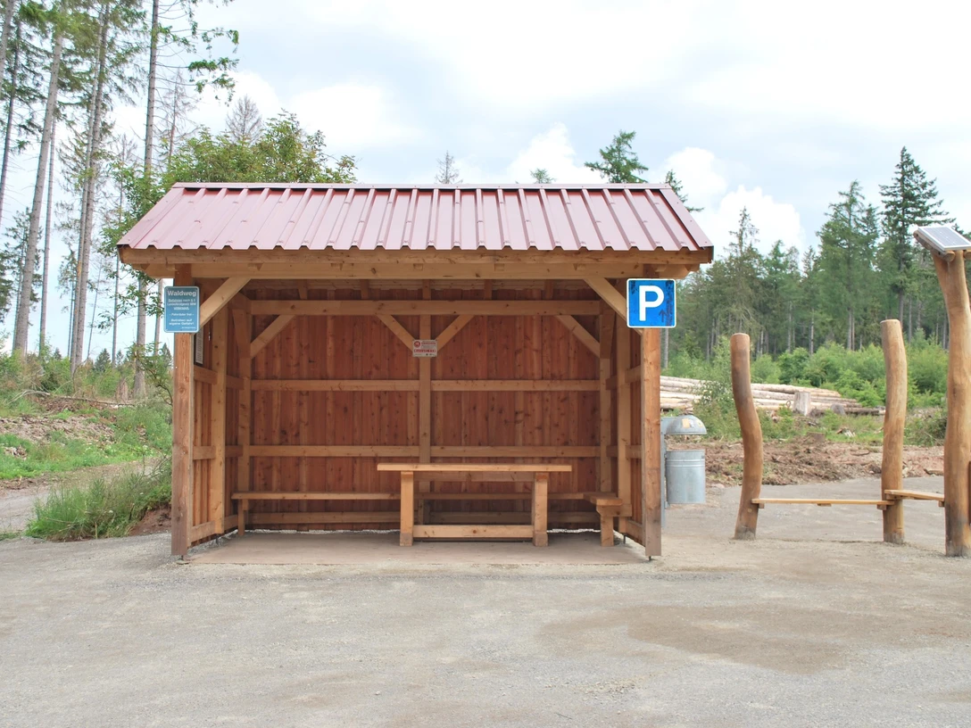 Hörstation 22 in Willebadessen Holzüberdachung mit Sitzbank im ländlichen Umfeld, Parkplatzsymbol und Informationstafeln daneben.