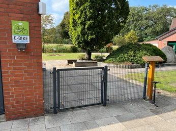 Hauskämpen Servicestation Eingangstor einer E-Bike Ladestation mit Gartenanlage und großzügiger Baumbestand im Hintergrund.