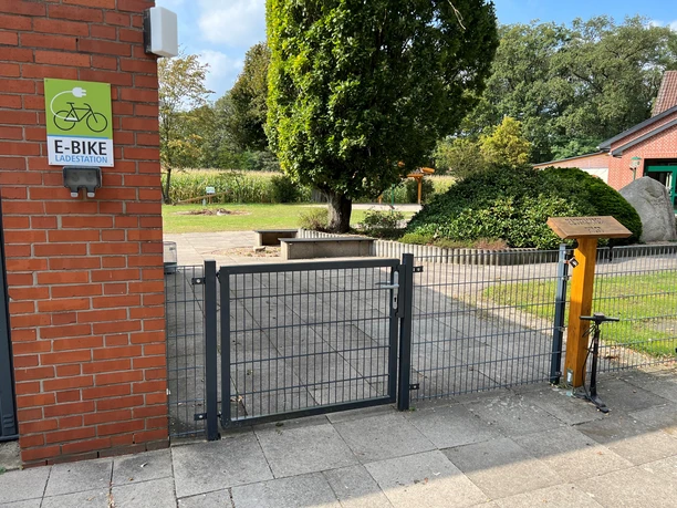 Hauskämpen Servicestation Eingangstor einer E-Bike Ladestation mit Gartenanlage und großzügiger Baumbestand im Hintergrund.