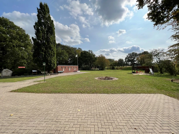 Ein gepflegter Rasenplatz mit gepflastertem Weg, Bäumen und einer Backsteinstruktur im Hintergrund.