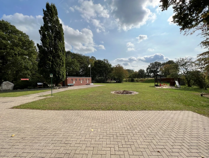 Hauskämpen Servicestation Ein gepflegter Rasenplatz mit gepflastertem Weg, Bäumen und einer Backsteinstruktur im Hintergrund.