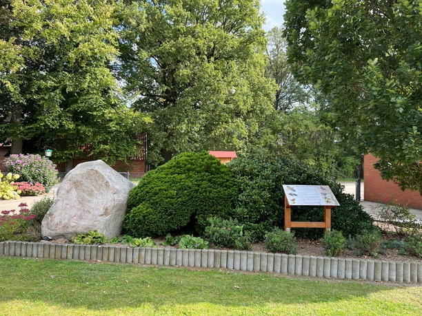 Grüne Vegetation mit einem großen Felsen und einer Infotafel, umgeben von Rasen und Bäumen.