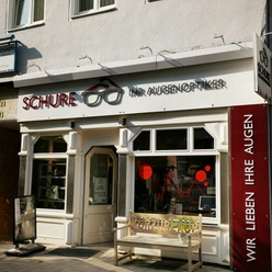 Schure.jpg Das Bild zeigt den Eingangsbereich des Geschäfts Schure. Vor dem Geschäft steht eine Bank
