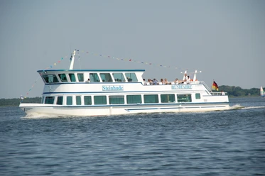 A white and blue passenger ship named Steinhude glides across a lake in the sunshine with people on board.
