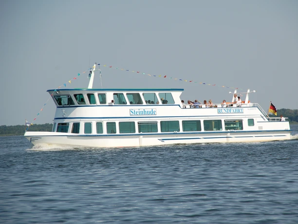 A white and blue passenger ship named Steinhude glides across a lake in the sunshine with people on board.