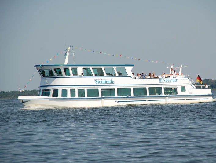 Ein weiß-blaues Fahrgastschiff namens Steinhude gleitet mit Menschen an Bord über einen See bei Sonnenschein.A white and blue passenger ship named Steinhude glides across a lake in the sunshine with people on board.Et hvidt og blåt passagerskib ved navn Steinhude glider over en sø i solskin med mennesker om bord.Een wit met blauw passagiersschip met de naam Steinhude glijdt over een meer in de zon met mensen aan boord.