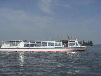 FGS Willkommen auf dem Steinhuder Meer Ein Ausflugsschiff mit Fähnchengirlanden fährt auf ruhigem Gewässer, im Hintergrund eine Baumlinie.An excursion boat with garlands of flags sails on calm waters, with a line of trees in the background.En udflugtsbåd med flagguirlander sejler på roligt vand med en række træer i baggrunden.Een excursieboot met vlaggenslingers vaart op kalm water, met een rij bomen op de achtergrond.