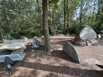 Findling "Alter Schwede aus dem Luk" Ein großer Findling, umgeben von Bäumen, neben Parkbänken und Picknicktisch, im lichten Wald.