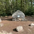 Findling "Alter Schwede aus dem Luk" Großer Findling im Wald, auf einem Steinsockel platziert, umgeben von Bäumen und einem Schild davor.