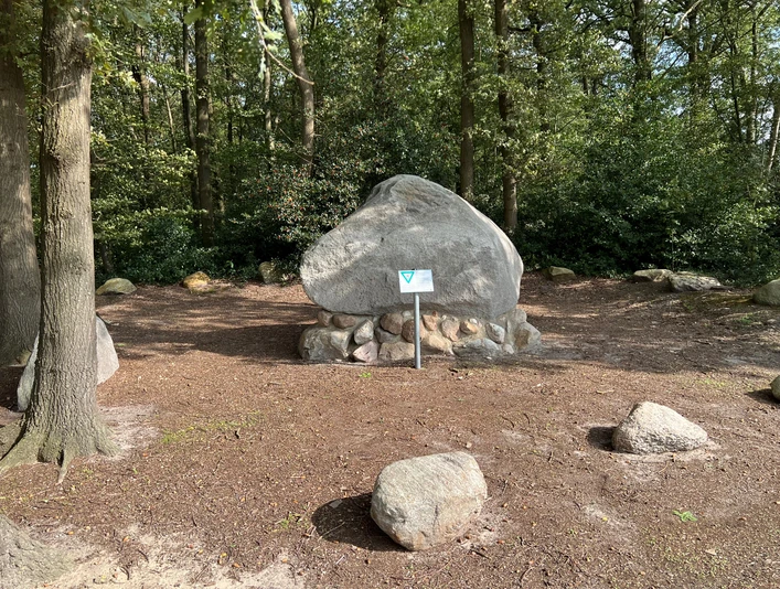 Findling "Alter Schwede aus dem Luk" Großer Findling im Wald, auf einem Steinsockel platziert, umgeben von Bäumen und einem Schild davor.